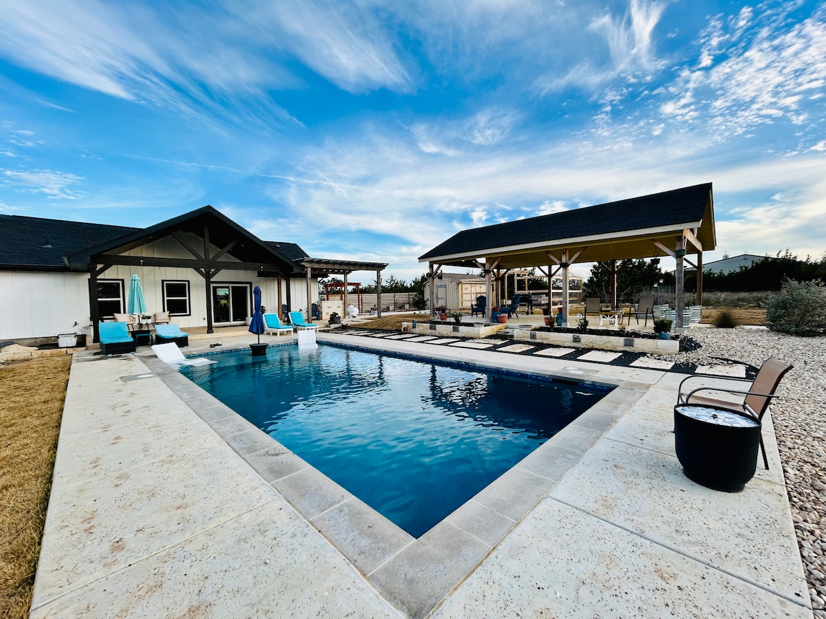 A resort-style pool area is showcased with a clear blue pool, surrounded by sun loungers and shaded seating. The property features a covered pavilion in the background, with a spacious lawn and a stone walkway leading toward it. Open skies enhance the serene atmosphere.
