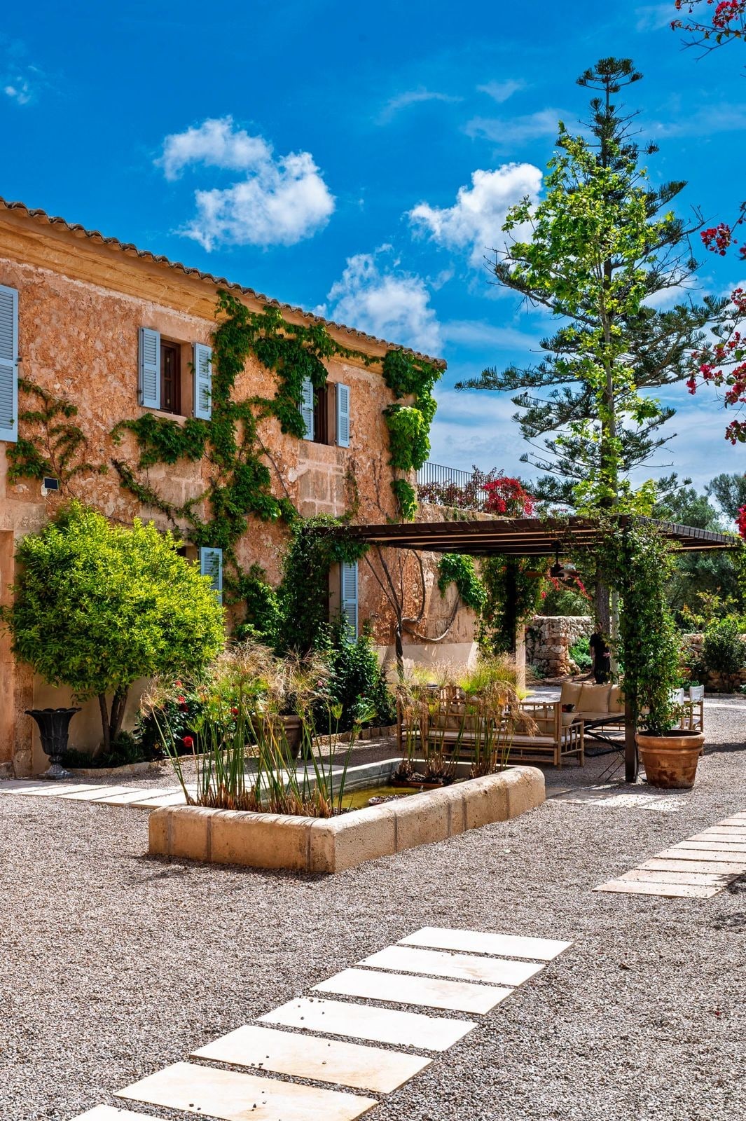 The exterior of a villa showcases a combination of natural and architectural elements. Lush greenery envelops the building, with flowering plants and trees adding vibrant touches. A gravel pathway leads toward a small pond, providing a serene focal point amidst the well-maintained landscape.