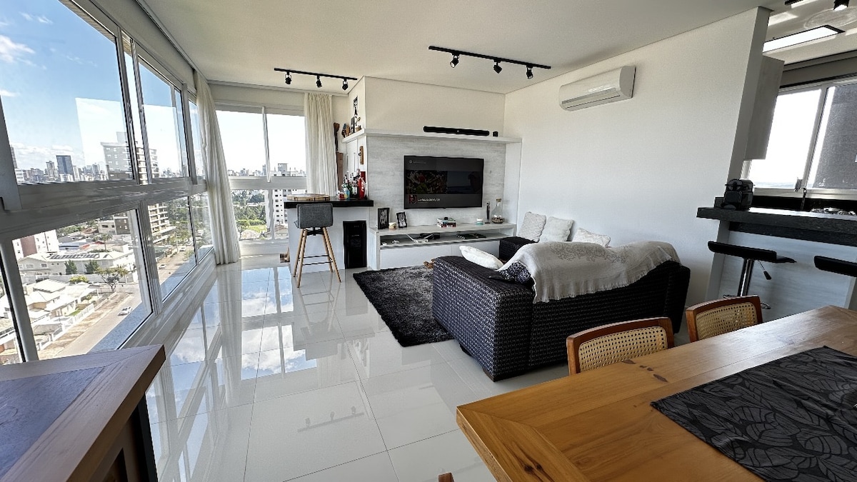 A spacious living area is showcased with large windows allowing natural light and city views. A comfortable couch rests on a dark rug, complemented by a sleek entertainment unit. A dining table with wooden chairs and a modern workspace are visible, creating a versatile setting.