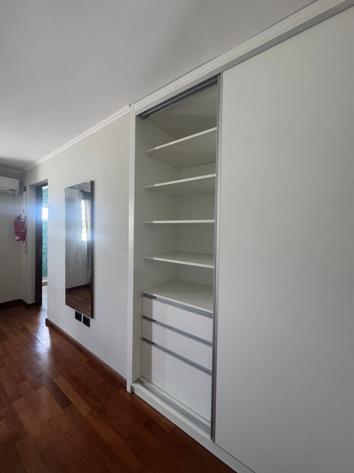 A spacious closet features open shelving and smooth sliding doors. Natural light enters through a nearby window, illuminating the area. A full-length mirror is mounted on the wall, providing a functional touch to the interior space.