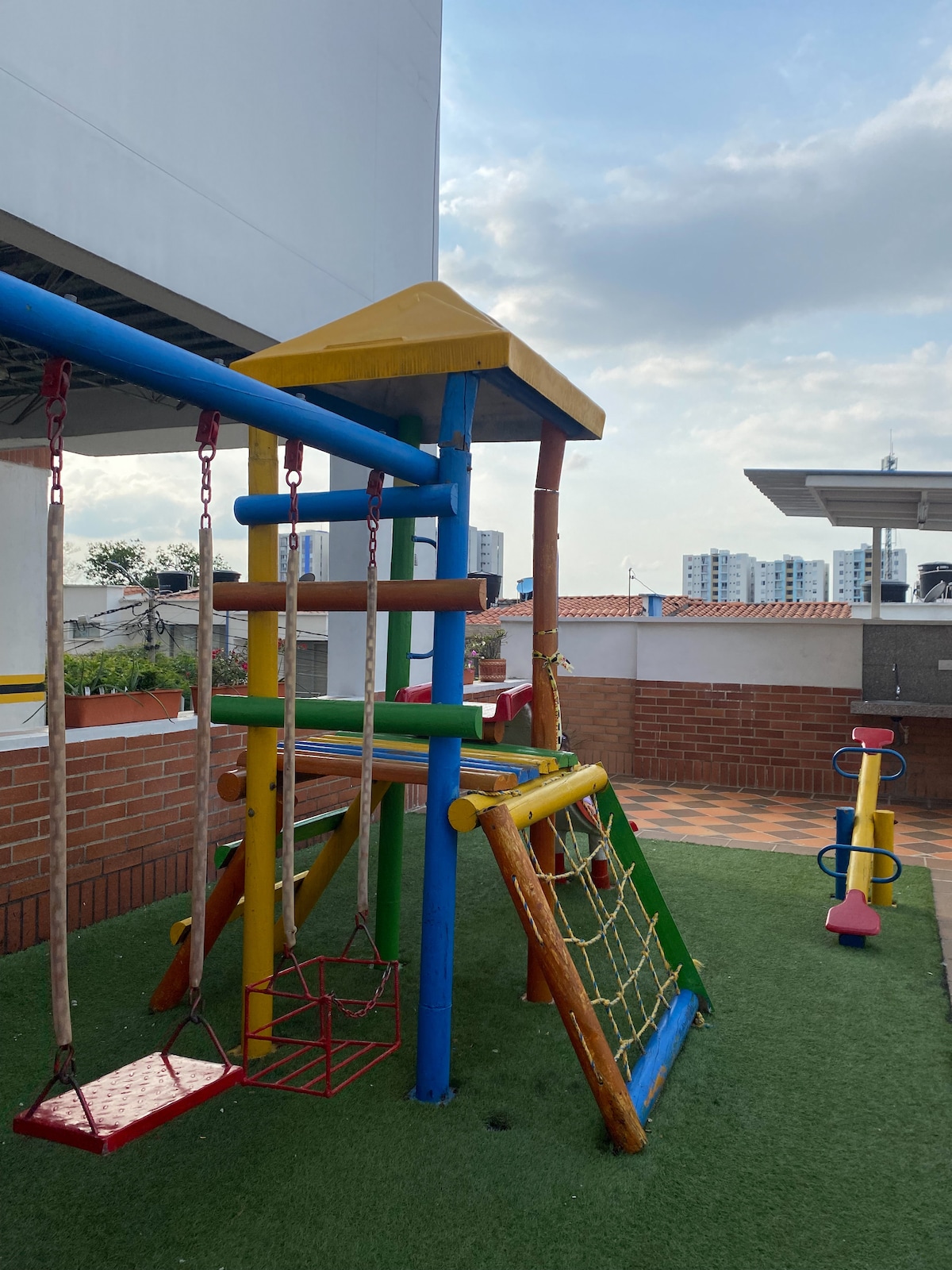 A colorful playground is featured in the outdoor area, comprising a play structure with slides and swings. Brightly painted elements, including red and yellow, add vibrancy. A soft green surface provides a safe play area, with an unobstructed view of the surrounding buildings.