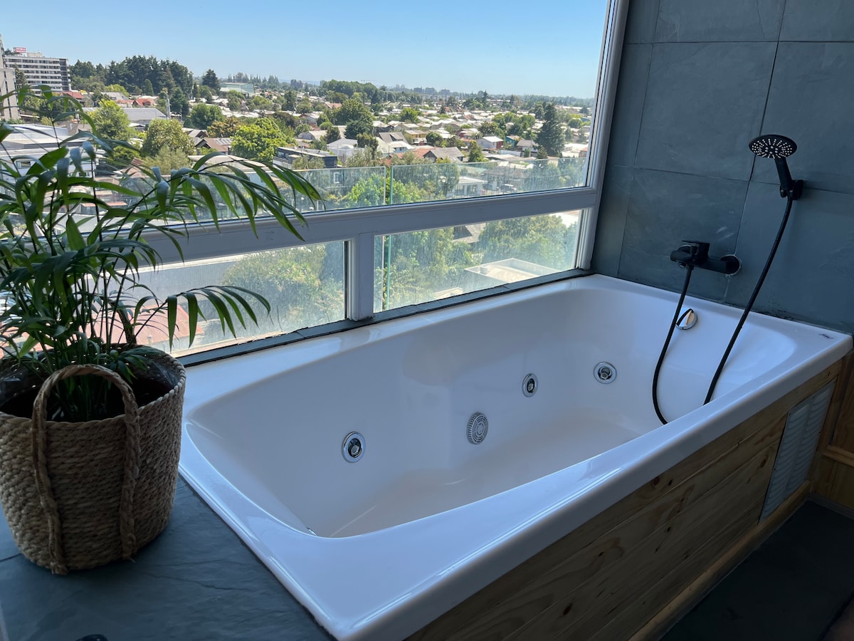 A spacious bathtub is positioned by a large window, offering a view of the surrounding landscape. Natural light illuminates the space, highlighting the sleek design of the tub and the modern shower fixture. A potted plant adds a touch of greenery to the setting.