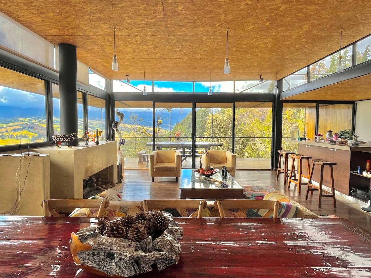 A spacious open-plan living area is presented, featuring large glass windows that frame scenic views of the mountains and valleys. Comfortable seating arrangements include soft armchairs and a coffee table, complemented by a rustic dining table. The natural light enhances the warm wood tones throughout.