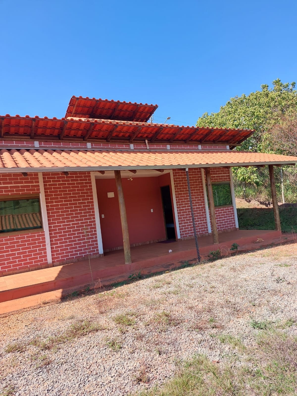 A charming exterior of a brick house is displayed, featuring a red-tiled roof and wooden posts supporting the overhang. Windows are covered with green shutters, allowing natural light to softly filter through. Surrounding greenery enhances the rural ambiance.