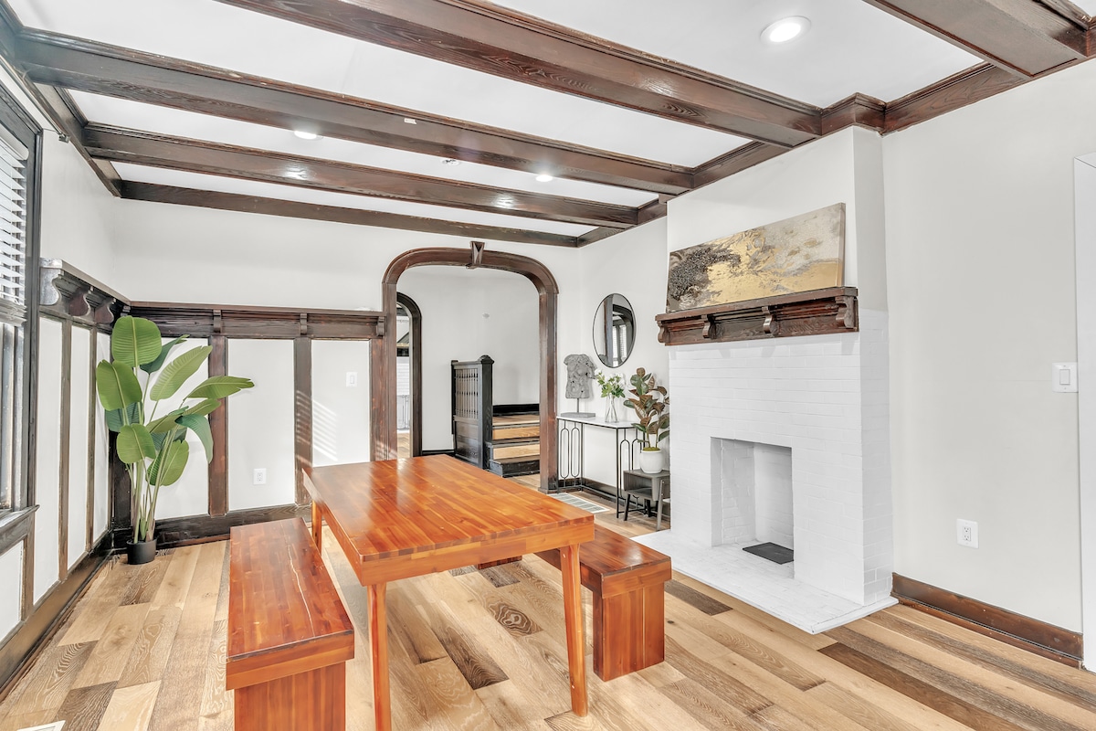 A spacious dining area is highlighted by a long wooden table with two matching benches. Dark wood beams adorn the ceiling, while a minimalist white fireplace serves as a central feature. Potted greenery adds a touch of nature to the room, complementing the warm wood flooring.