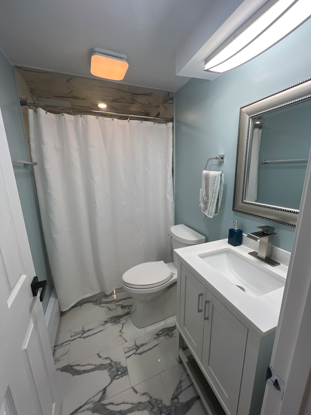 A newly renovated bathroom features a walk-in shower with a white curtain and a sleek vanity with a modern sink. Light blue walls and marble-patterned flooring provide a clean, refreshing look. A mirror above the sink reflects the gentle illumination from the ceiling light.