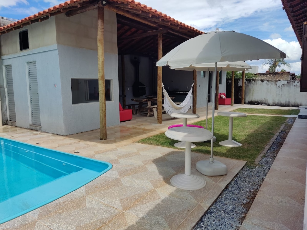 A relaxed outdoor area is showcased, featuring a turquoise pool alongside a shaded umbrella table. A hammock is visible, with a grassy patch and patterned stone tiles surrounding the space. The building structure features wooden beams and stone walls, providing a perfect setting for leisure.