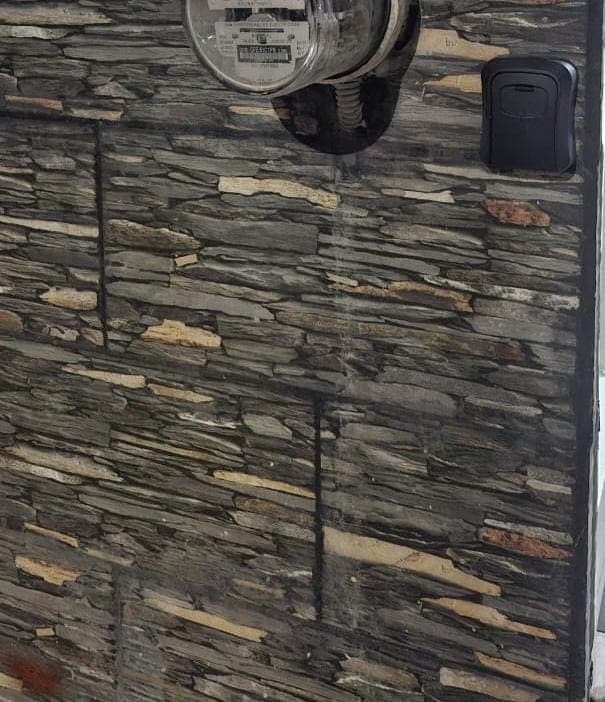 A textured wall features natural stone tiles in varying shades of gray and brown, creating a layered appearance. A glass container is mounted on the wall, and a small black device is attached beside it. The setting conveys a clean and understated aesthetic.