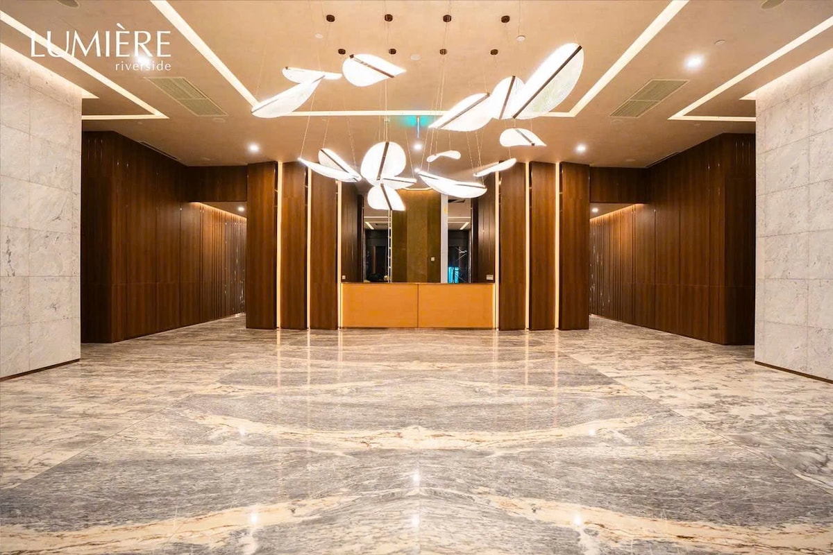 An elegantly designed lobby features a spacious marble floor, highlighted by an intricate lighting installation that resembles floating leaves. Rich wooden paneling surrounds the area, creating a warm and modern aesthetic. A reception desk is positioned at the end of the lobby, inviting guests.