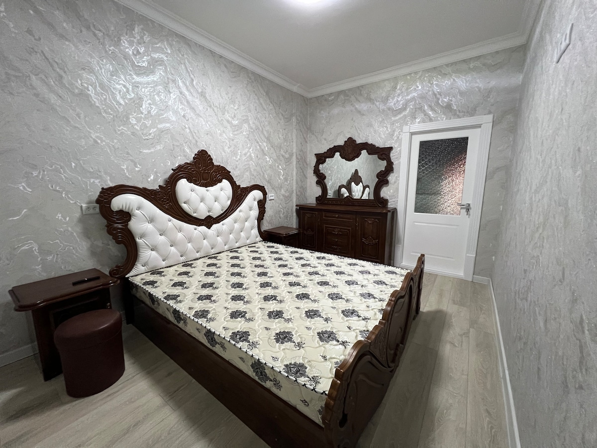 A spacious bedroom features an ornate wooden bed with a padded headboard. A matching wooden dresser and mirror complement the rich decor. Light-colored walls and flooring enhance the room's brightness, while a small round stool is positioned beside the bed.