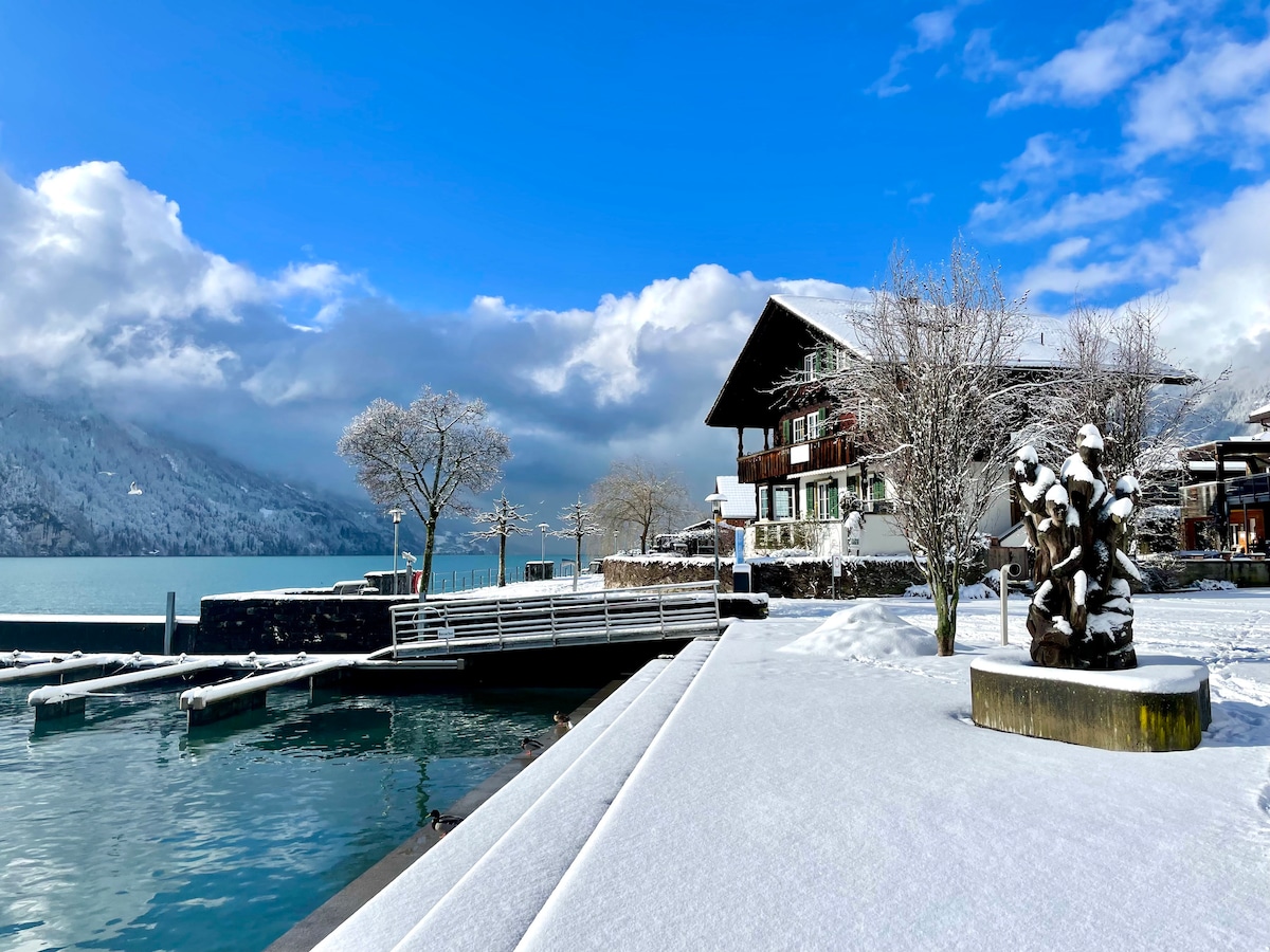 A winter scene showcases a snow-covered landscape featuring a tranquil lake. A traditional Swiss chalet stands in the background, while a modern sculpture and trees line the pathway leading to the water's edge. The sky is partly cloudy, creating a serene atmosphere.