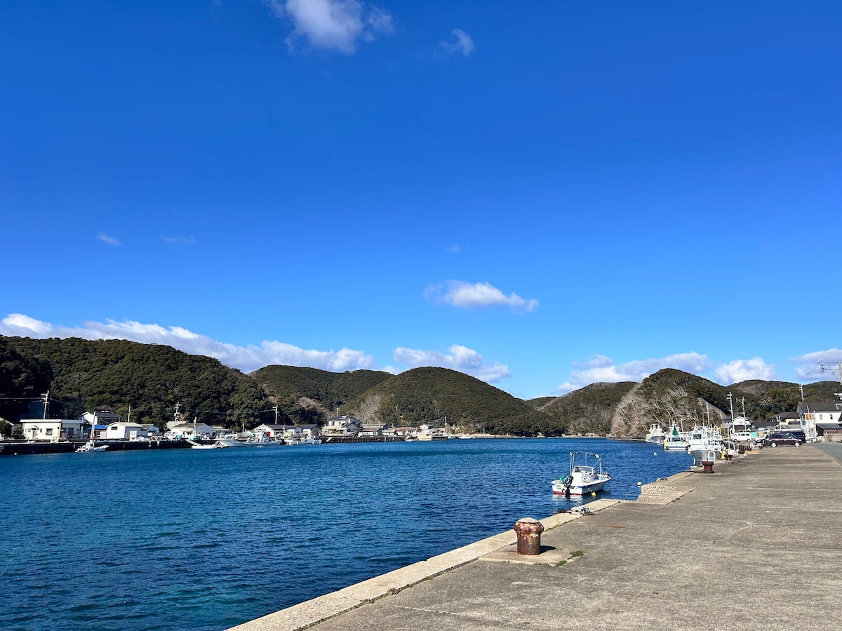 A serene harbor is depicted with calm blue waters lined by a concrete pier. Small boats are anchored, and quaint buildings can be seen along the waterfront. Rolling green hills rise in the background under a clear blue sky with a few fluffy clouds.