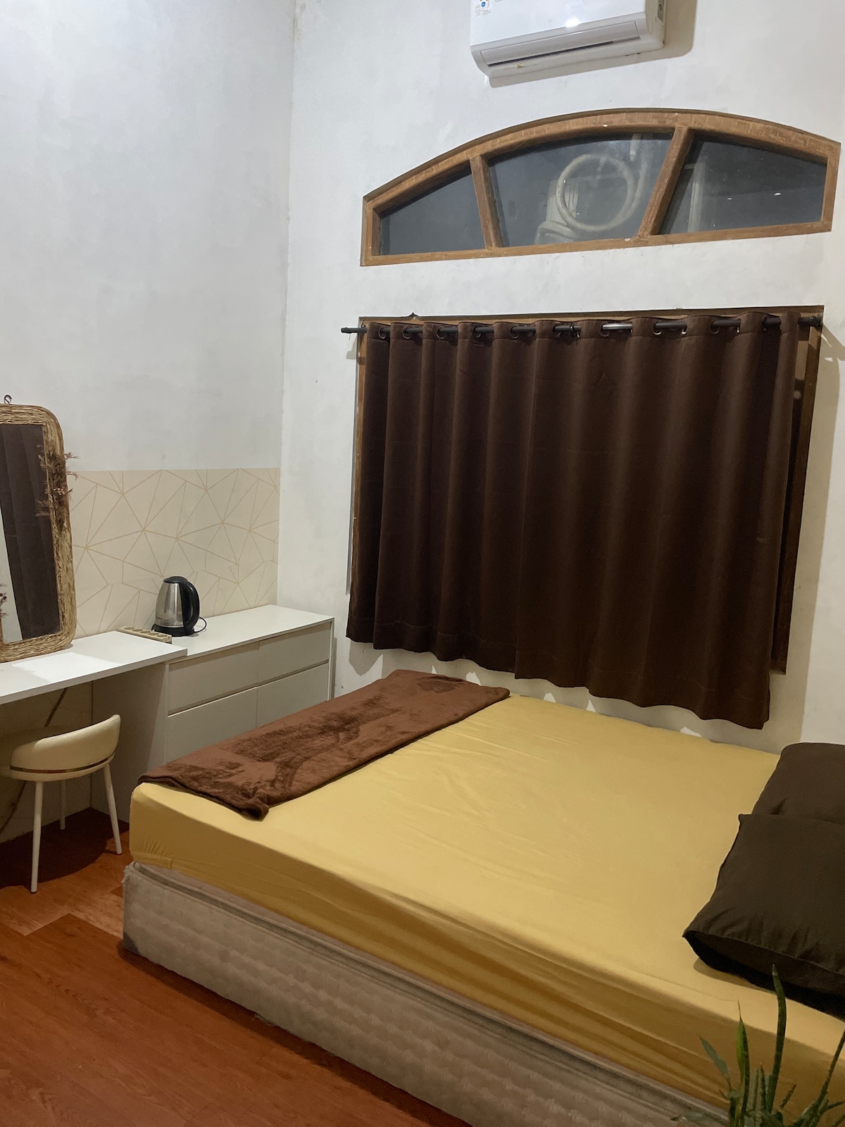 A compact sleeping area is displayed with a spacious bed covered in a neutral-colored blanket. A small desk and chair are positioned next to an electric kettle. Dark curtains frame a window, allowing soft light to enter the room.