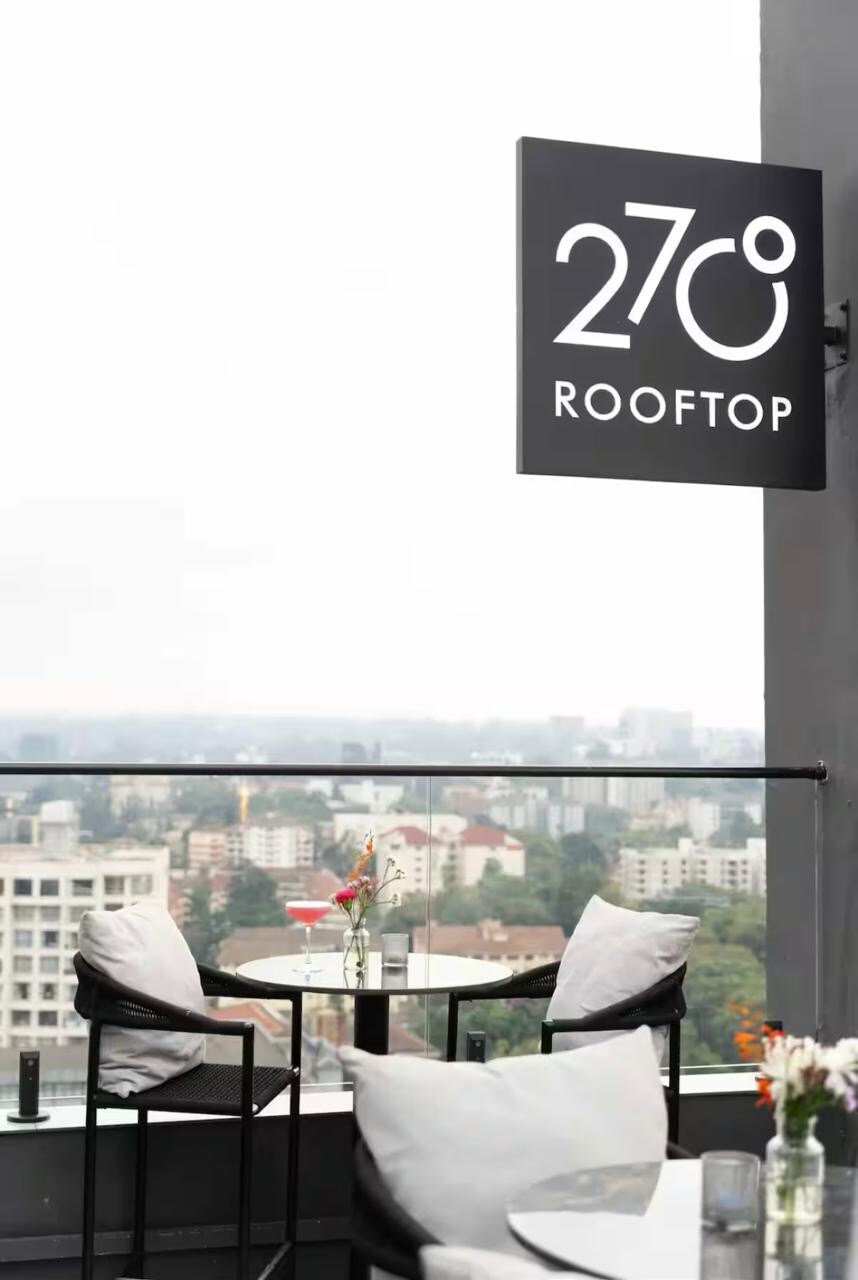 A rooftop space is captured with a clear sign displaying '270° ROOFTOP.' Two modern, round tables with dark metal frames are positioned on a balcony, overlooking a cityscape. Small vases with flowers are placed on the tables, and a glass railing enhances the view.