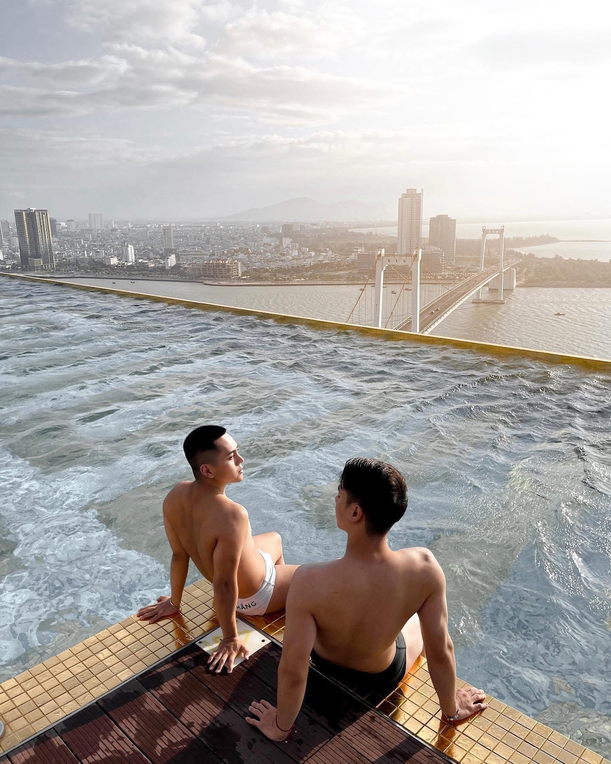 An infinity pool extends towards the horizon, offering breathtaking views of the city skyline and a nearby bridge. The pool's edge creates a seamless transition between water and skyline. Soft sunlight illuminates the scene, enhancing the tranquil atmosphere of the space.