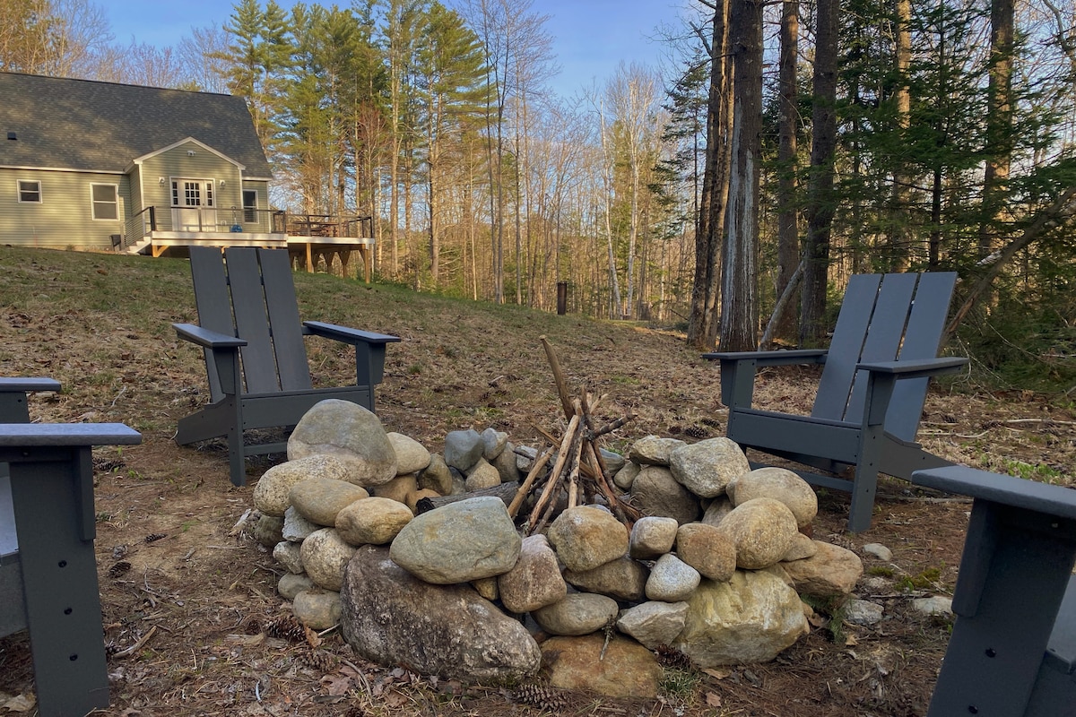 A cozy fire pit area is surrounded by grey Adirondack chairs, positioned on natural stone. The fire pit, constructed with smooth stones, is ready for outdoor gatherings. In the background, a spacious home is nestled among tall trees, creating a serene, wooded setting.
