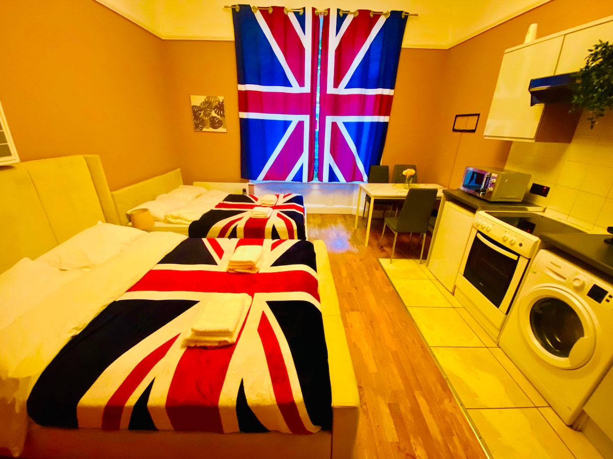 A light-filled studio features two spacious beds adorned with Union Jack blankets. A kitchenette is equipped with essential appliances, including a microwave and washing machine. The wall is decorated with a large British flag curtain, complementing the warm-toned walls and wooden flooring.