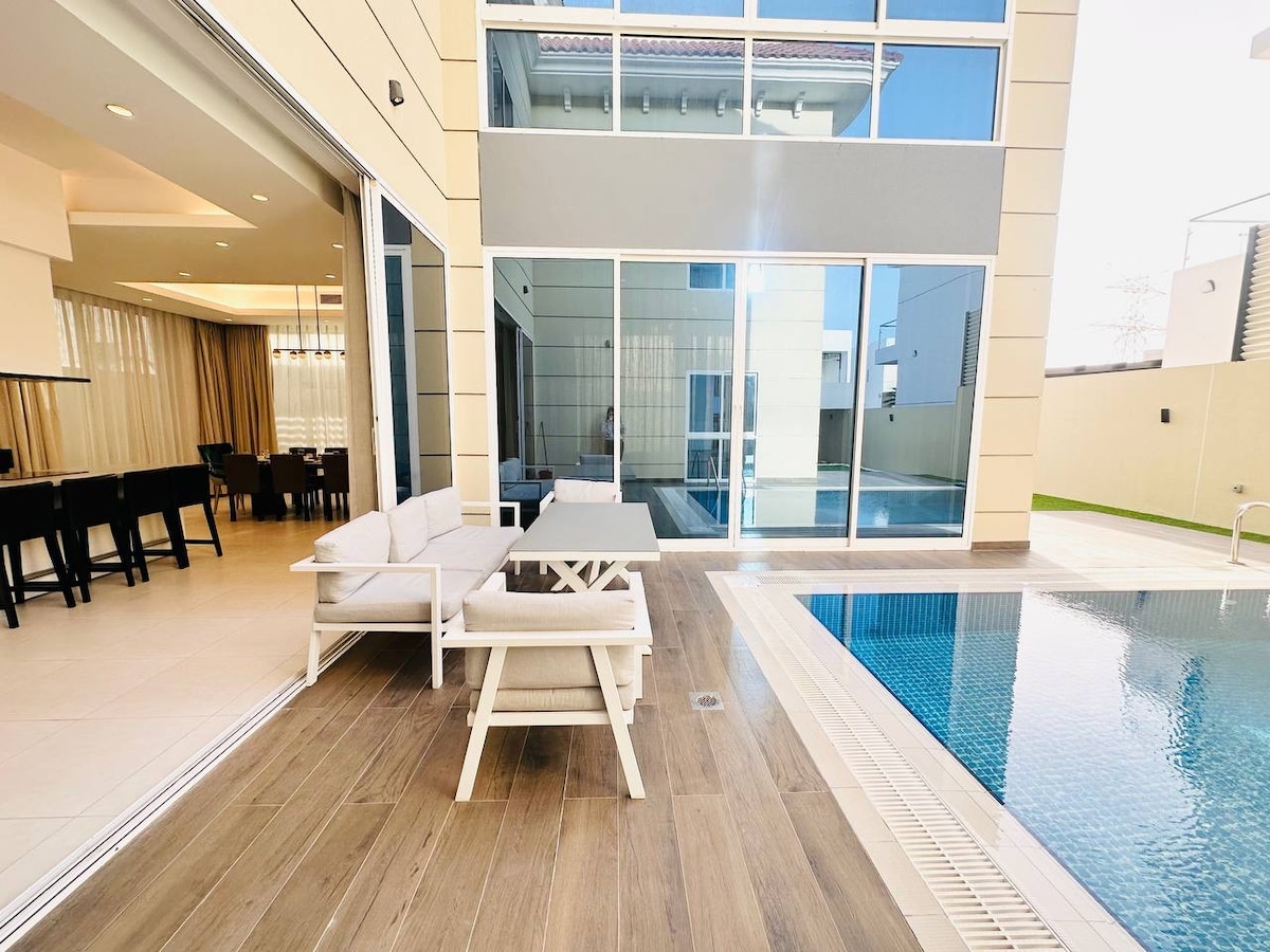 An outdoor seating area features a white table and matching chairs, positioned on a wooden deck beside a pool. The spacious area is surrounded by tall glass doors leading to a well-lit indoor space, creating an open and airy feel.