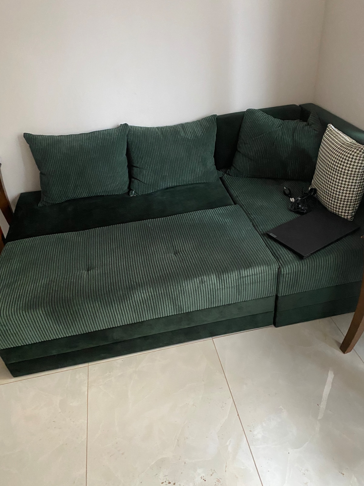 A modern green sectional sofa is featured with plush cushions in a ribbed texture. The seating area is complemented by a neutral-patterned throw pillow and a small laptop resting on the arm, creating a cozy spot for relaxation or work.