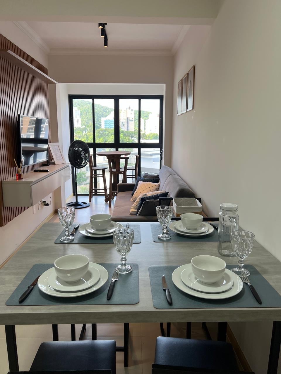 A dining table set for four is positioned in the foreground, featuring plates, glasses, and cutlery. A comfortable sofa is situated along the back wall, adjacent to a small dining area with stool seating. A fan is visible beside the television, with large windows allowing natural light.