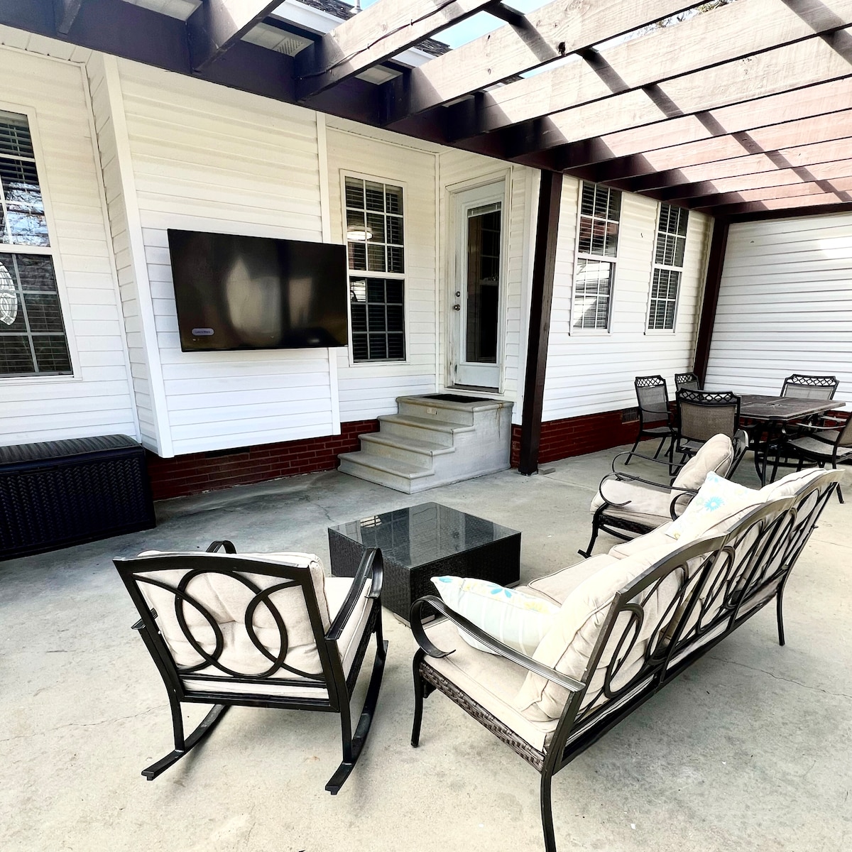 An outdoor entertainment area features a covered patio with a seating arrangement that includes two cushioned chairs and a sofa surrounding a coffee table. A mounted TV is visible on the wall, enhancing the space for relaxation and gatherings.