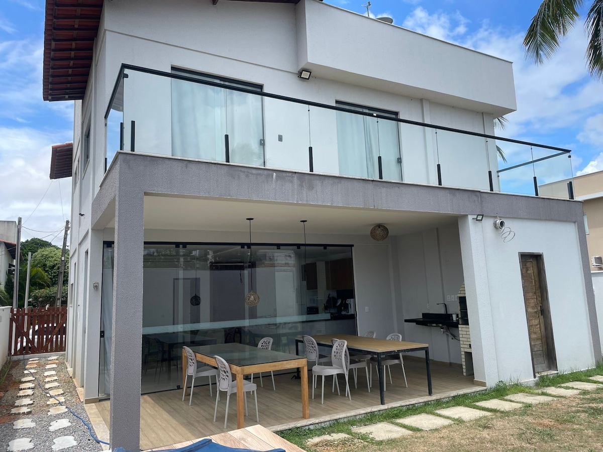 A modern two-story house is shown featuring a spacious balcony with glass railing. The ground level includes a covered outdoor dining area with a large table and several chairs, alongside a built-in barbecue. The well-maintained lawn adds to the inviting outdoor space.