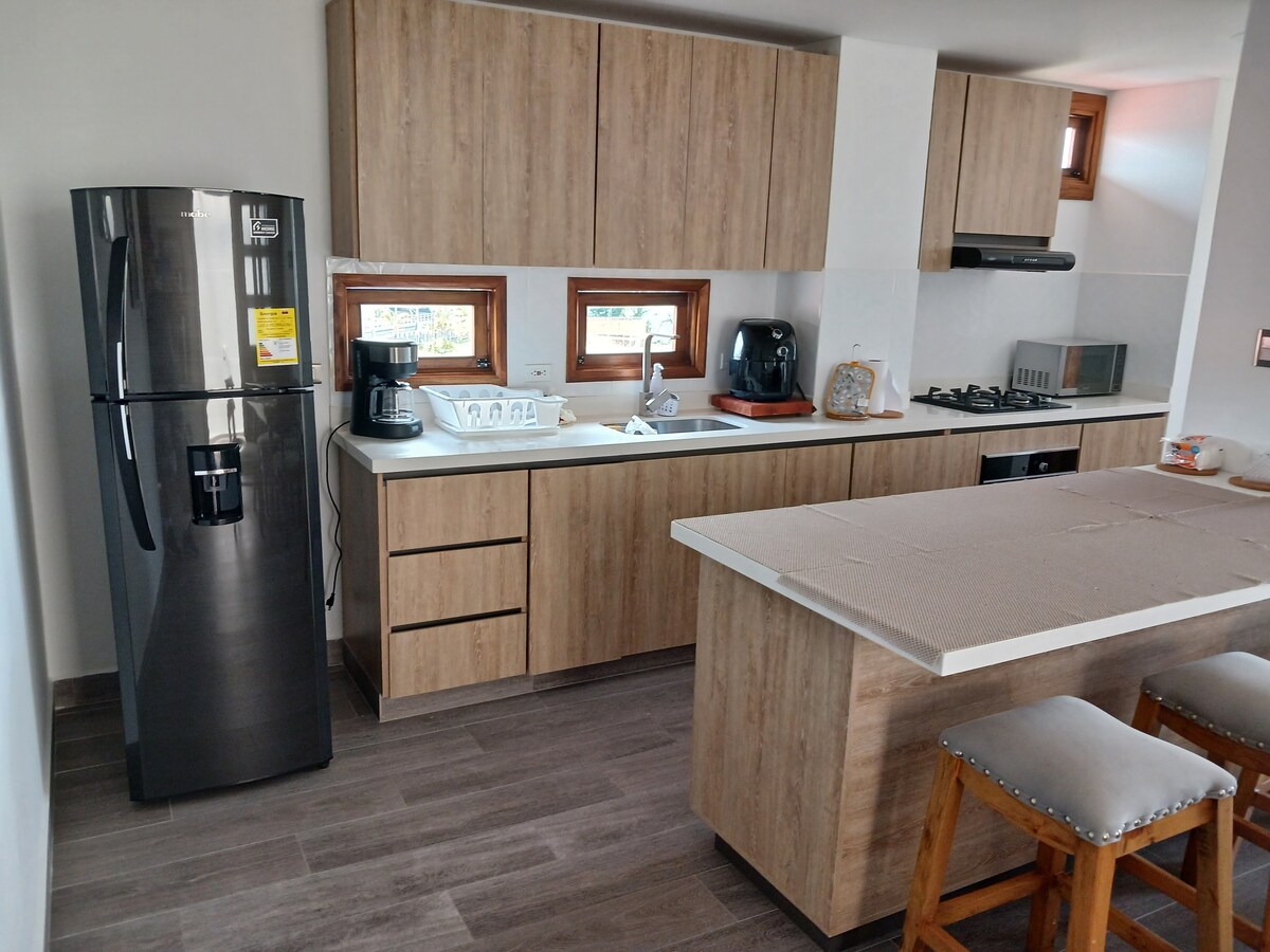 A well-equipped kitchen features wooden cabinetry and modern appliances. A black refrigerator stands next to a sleek countertop with a sink. Two bar stools are positioned at an island, and natural light enters through multiple windows.