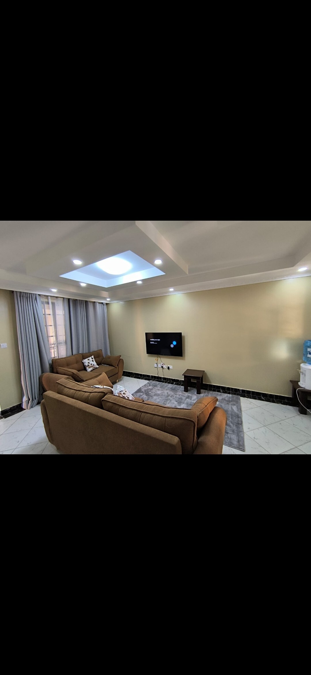 A cozy living area features a light brown sectional sofa centered around a wall-mounted TV. Soft lighting is provided by recessed ceiling fixtures, and large grey curtains frame the window. A small side table is positioned beside the sofa, contributing to a comfortable environment.