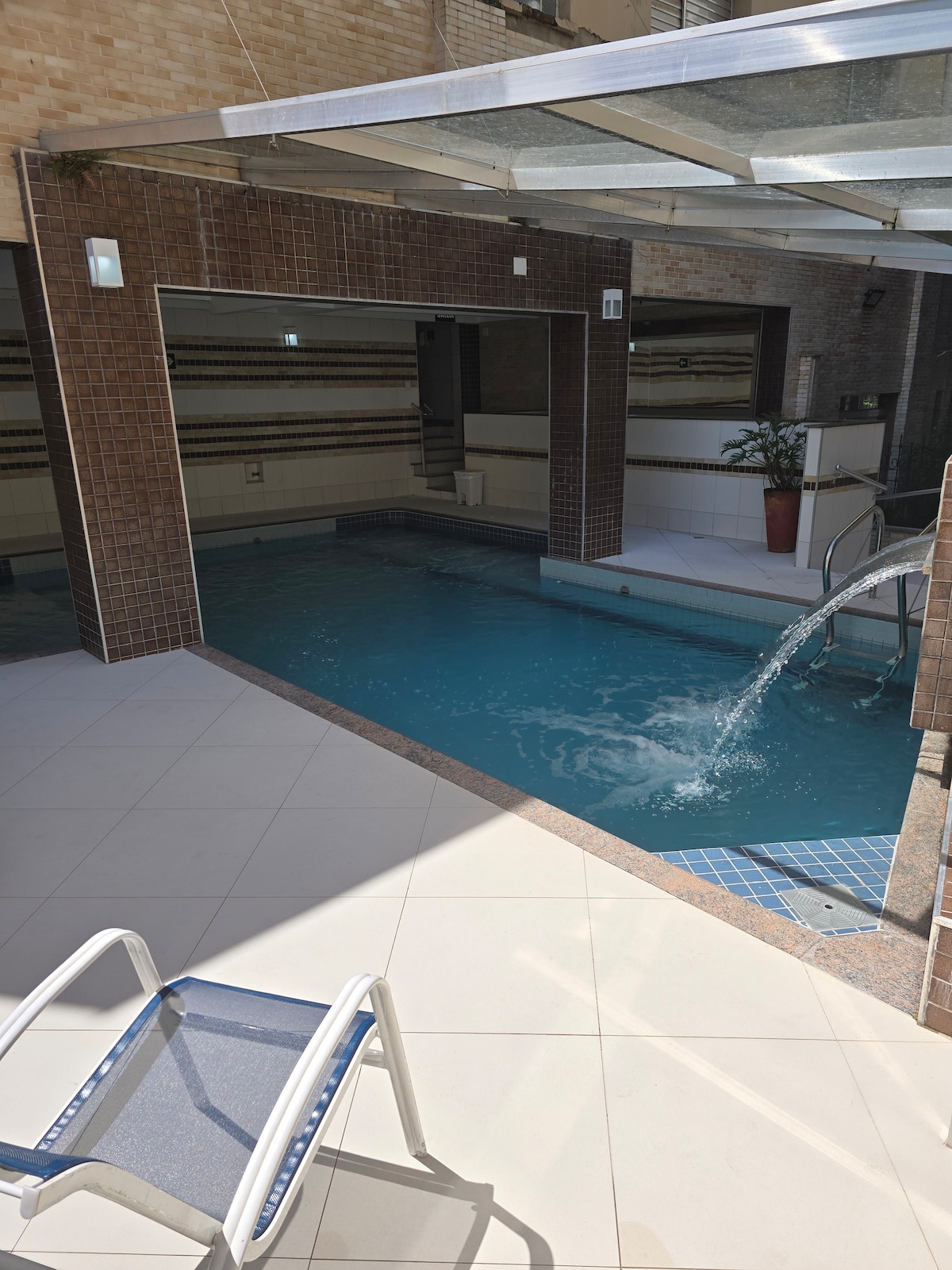 A serene indoor pool area features clear water with a gentle waterfall. Surrounding walls are tiled with neutral tones, and potted plants add a touch of greenery. A sturdy white lounge chair is positioned nearby on the light-colored tiled floor.