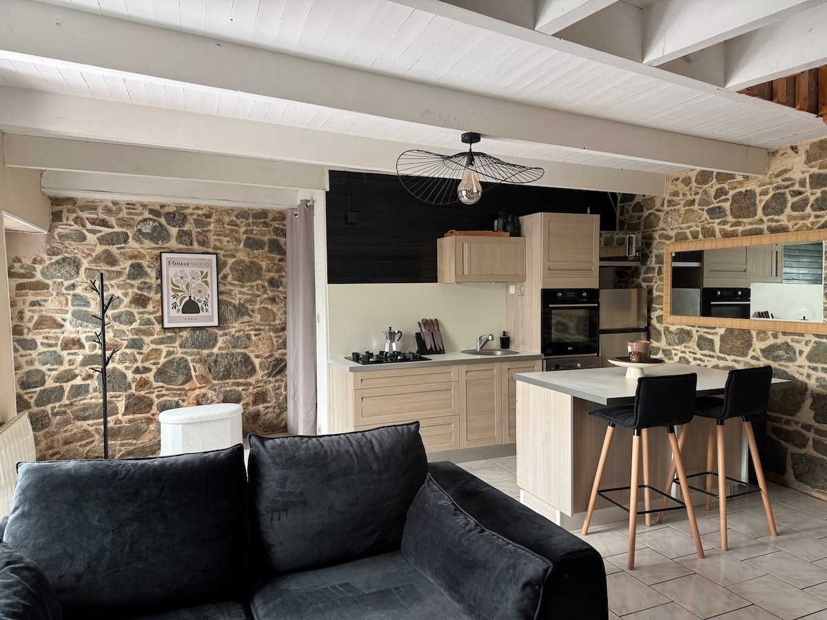 A spacious kitchen and living area feature exposed stone walls and wooden beams. Modern appliances are integrated into a stylish kitchen with light cabinetry. A comfortable black sofa is positioned nearby, accompanied by bar-style chairs at a counter, offering a welcoming space for relaxation and dining.