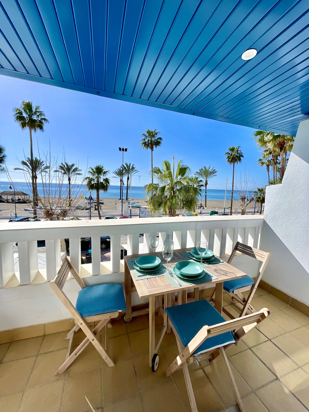 A terrace is pictured with a table set for four, featuring blue and beige tones. The space is framed by a white railing, with views of palm trees and the beach in the background. The blue ceiling adds a bright touch, completing the seaside ambiance.