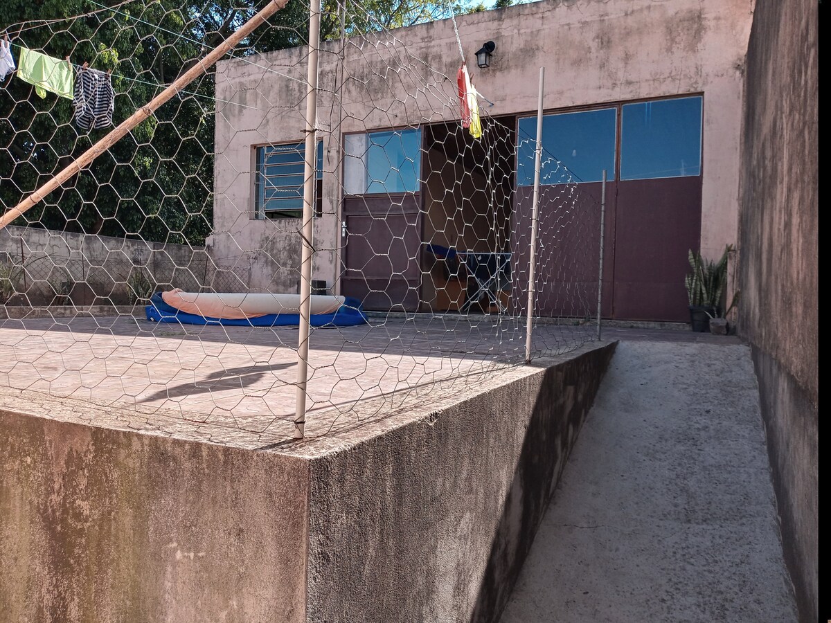 An exterior view showcases a fenced area with a concrete pathway leading up to a building. The entrance features large doors, and a few colorful items hang on a line. A blue inflatable pool rests in the fenced area, complemented by greenery in pots.