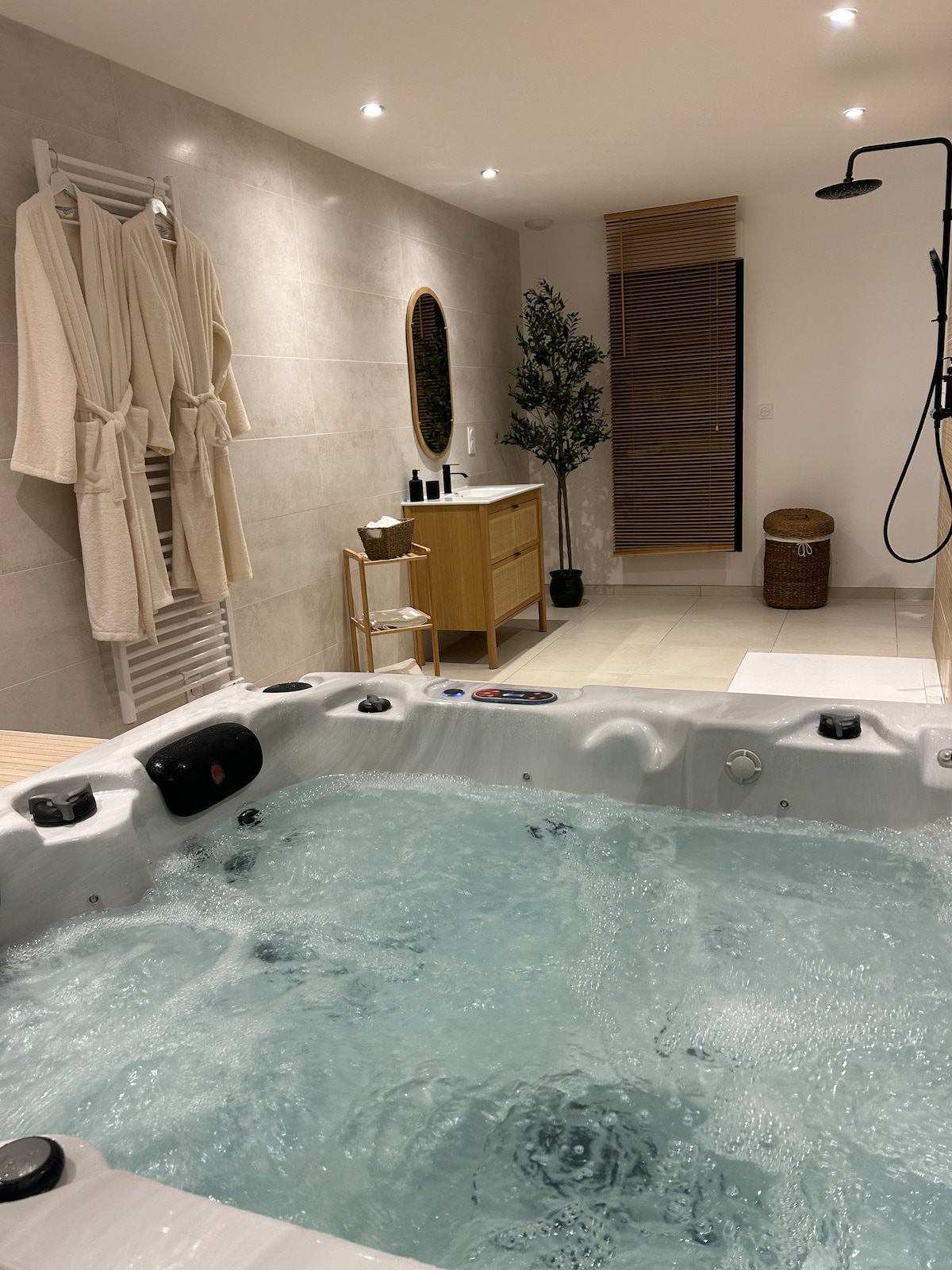 A spacious jacuzzi filled with bubbling water is featured in a well-lit spa area. Soft lighting highlights the neutral-toned walls and wooden elements, including a vanity and a woven basket. Luxurious robes hang on a rack, and a plant adds a touch of greenery.