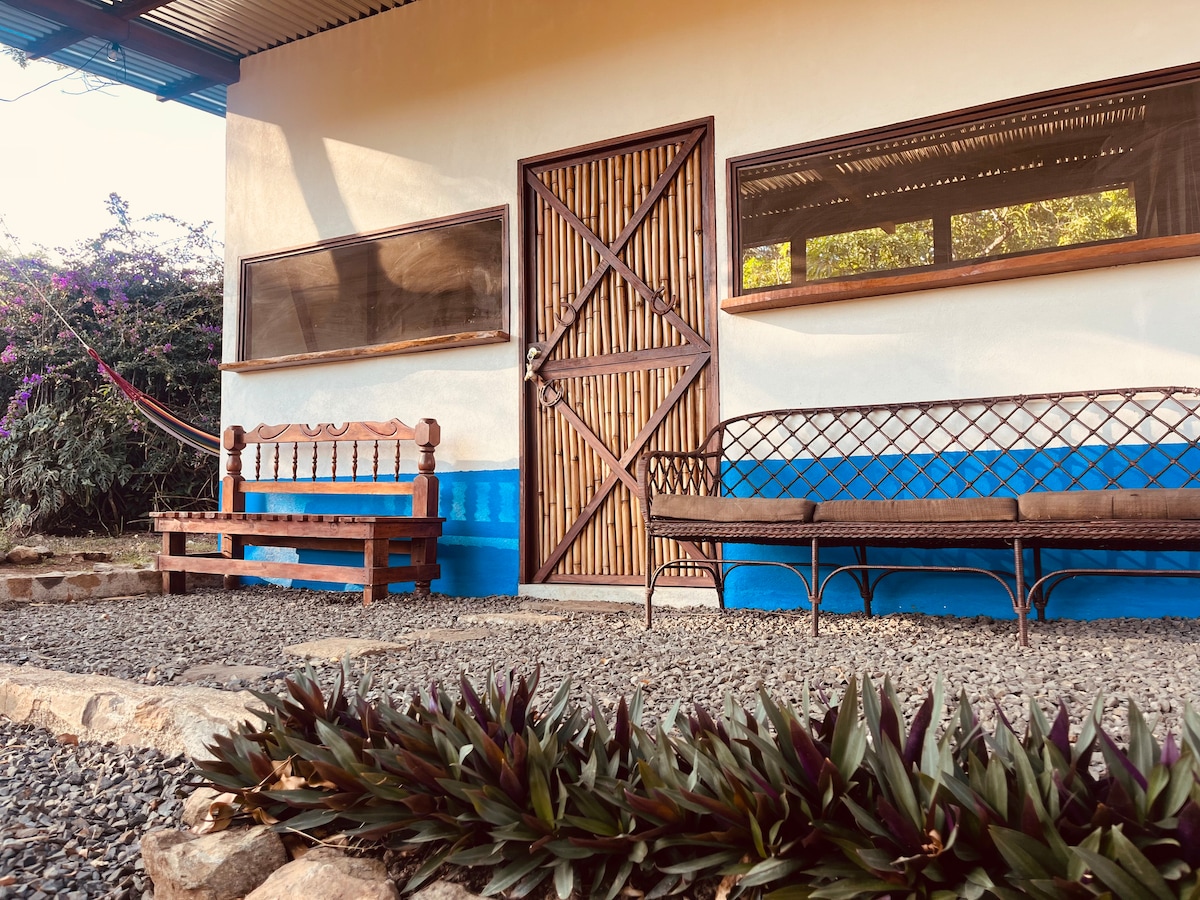 The exterior of the cottage features a wooden door framed by large windows. A wooden bench and two metal benches are placed on a gravel surface, inviting relaxation. Tropical plants adorn the area, enhancing the outdoor space's natural charm.