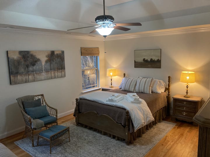 Dormitorio principal que muestra una cama tamaño queen lujosamente equipada con lámparas de lectura y una sala de estar. Las ventanas dan al patio trasero privado con bosque.