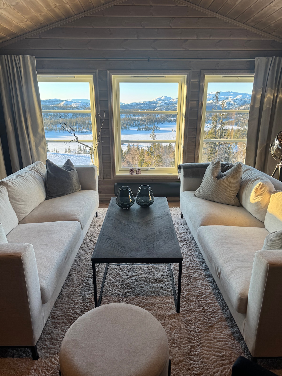 Two light-colored sofas face each other across a dark wooden coffee table in a cozy corner. Large windows provide a panoramic view of a snowy landscape, with mountains in the distance and a serene lake visible, creating a tranquil ambiance.