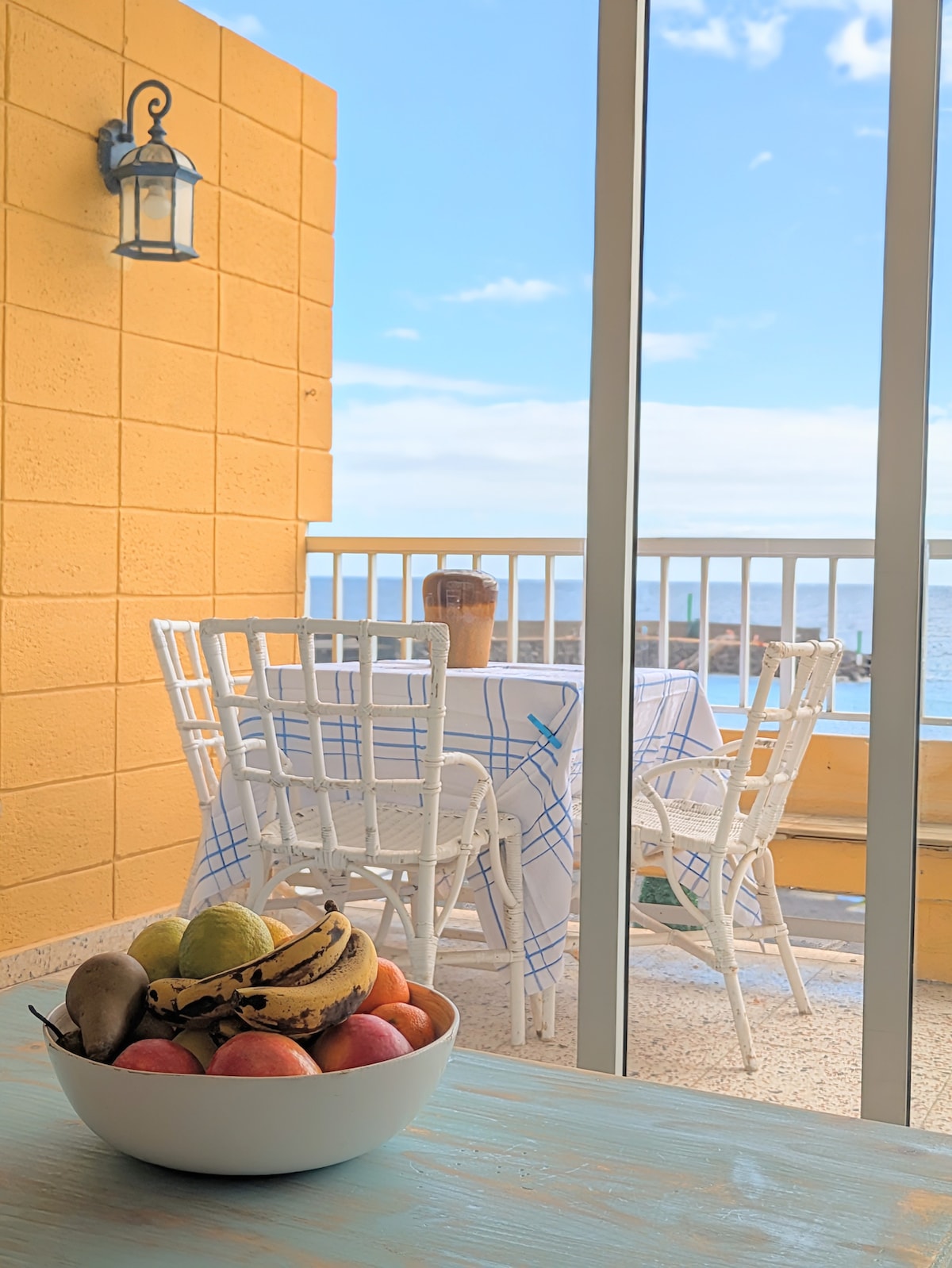 A bright balcony with a table and chairs overlooks the ocean. A bowl of fresh fruits sits on a nearby table, and light pours in through the sliding glass doors, enhancing the open and airy feel of the space.