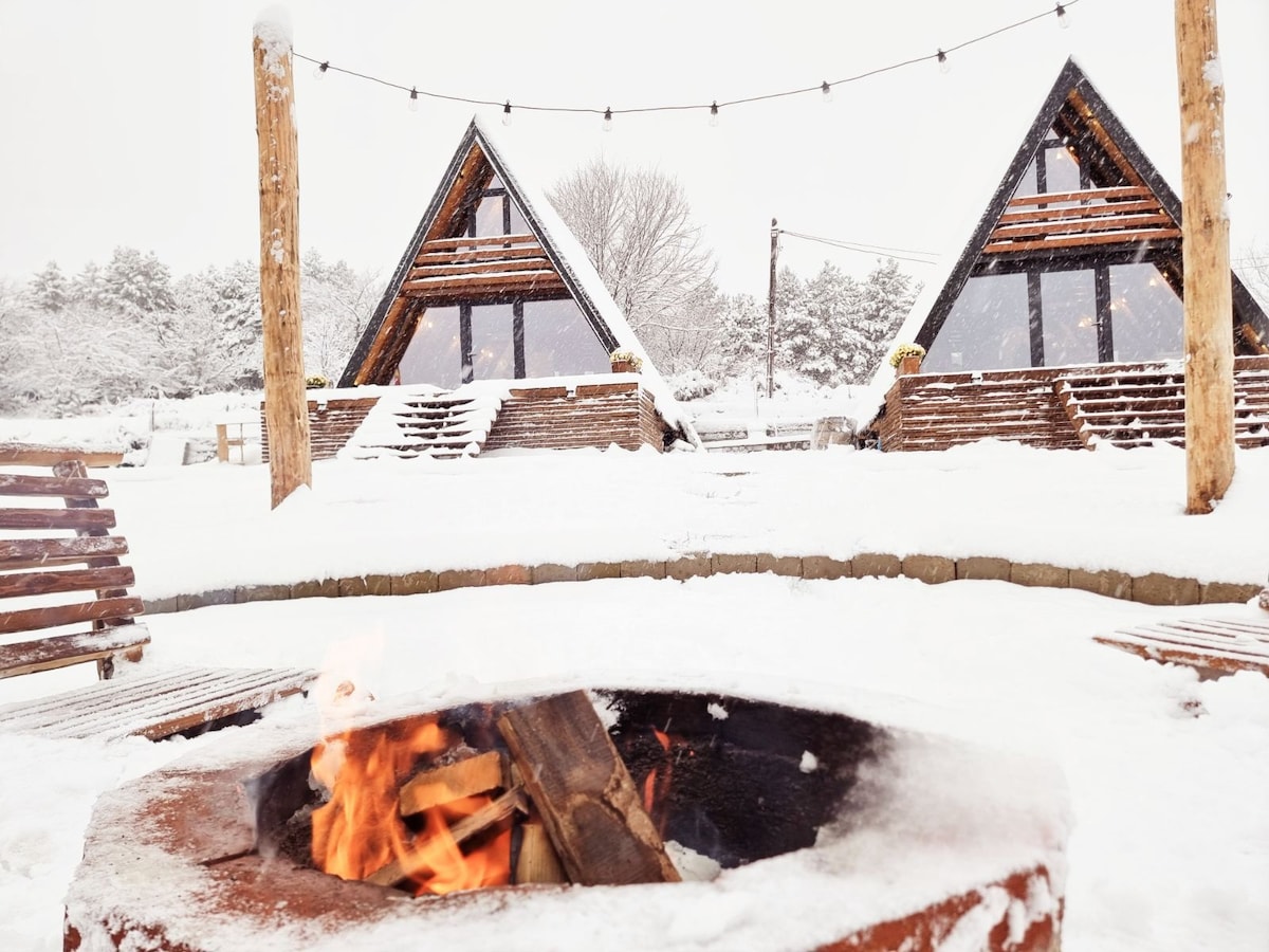 Two A-frame cabins are visible in the background, surrounded by a snowy landscape. A cozy fire pit sits in the foreground, with flames gently flickering among stacked firewood. String lights hang above, adding a warm ambiance to the winter scene.