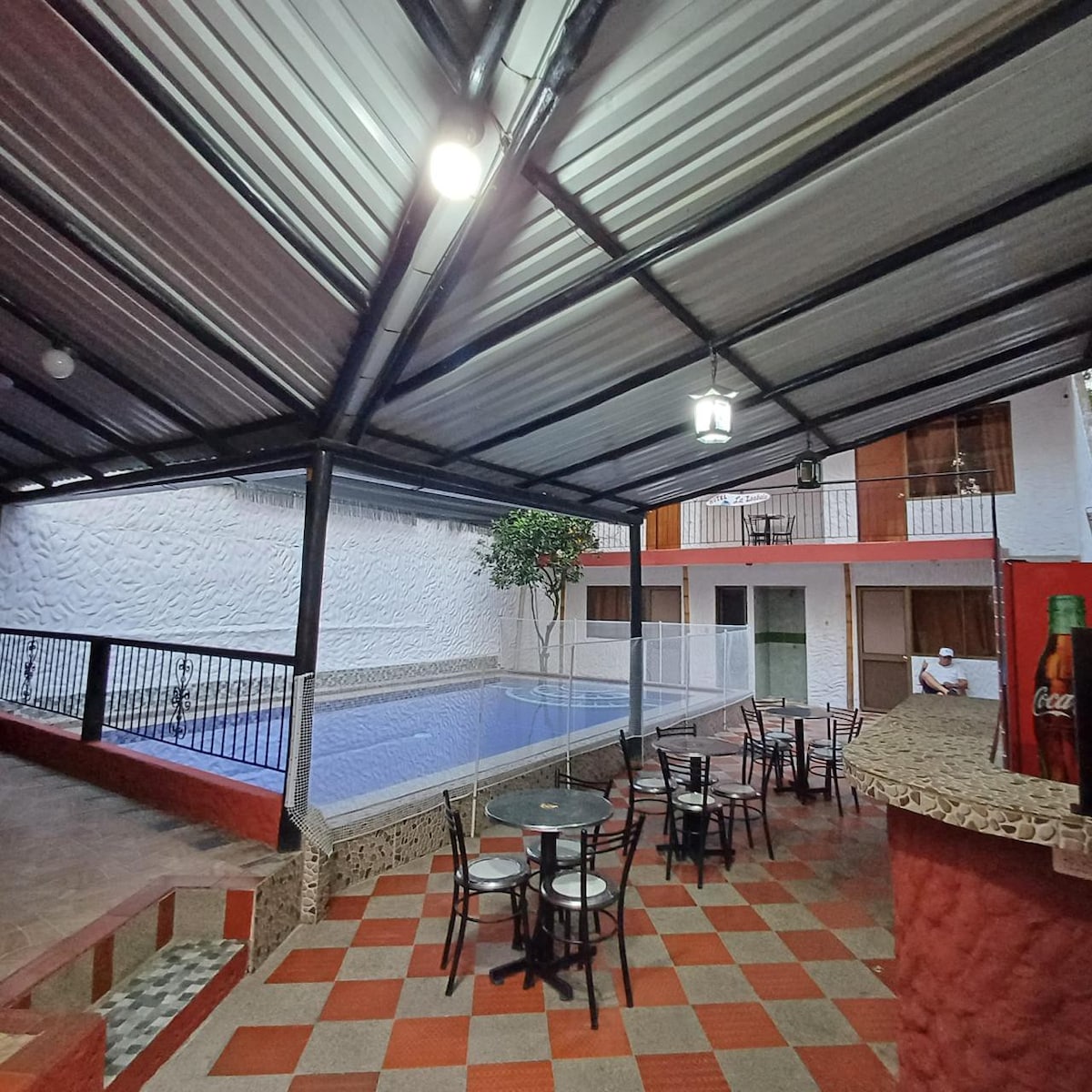 A covered pool area features a spacious swimming pool surrounded by a protective fence. Small round tables with chairs are arranged nearby on a patterned tiled floor, contributing to a relaxed ambiance. Light filters in from the roof, illuminating the inviting space.
