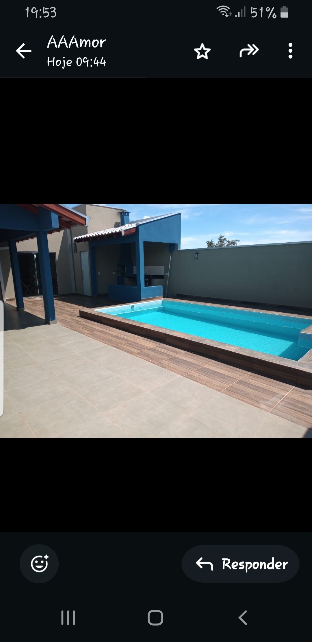 A rectangular swimming pool features clear turquoise water, surrounded by a tiled deck. Sunlight reflects off the water, while a covered patio area with pillars stands adjacent, providing shaded seating. The backdrop includes a neutral-colored wall and blue accents on the building structure.