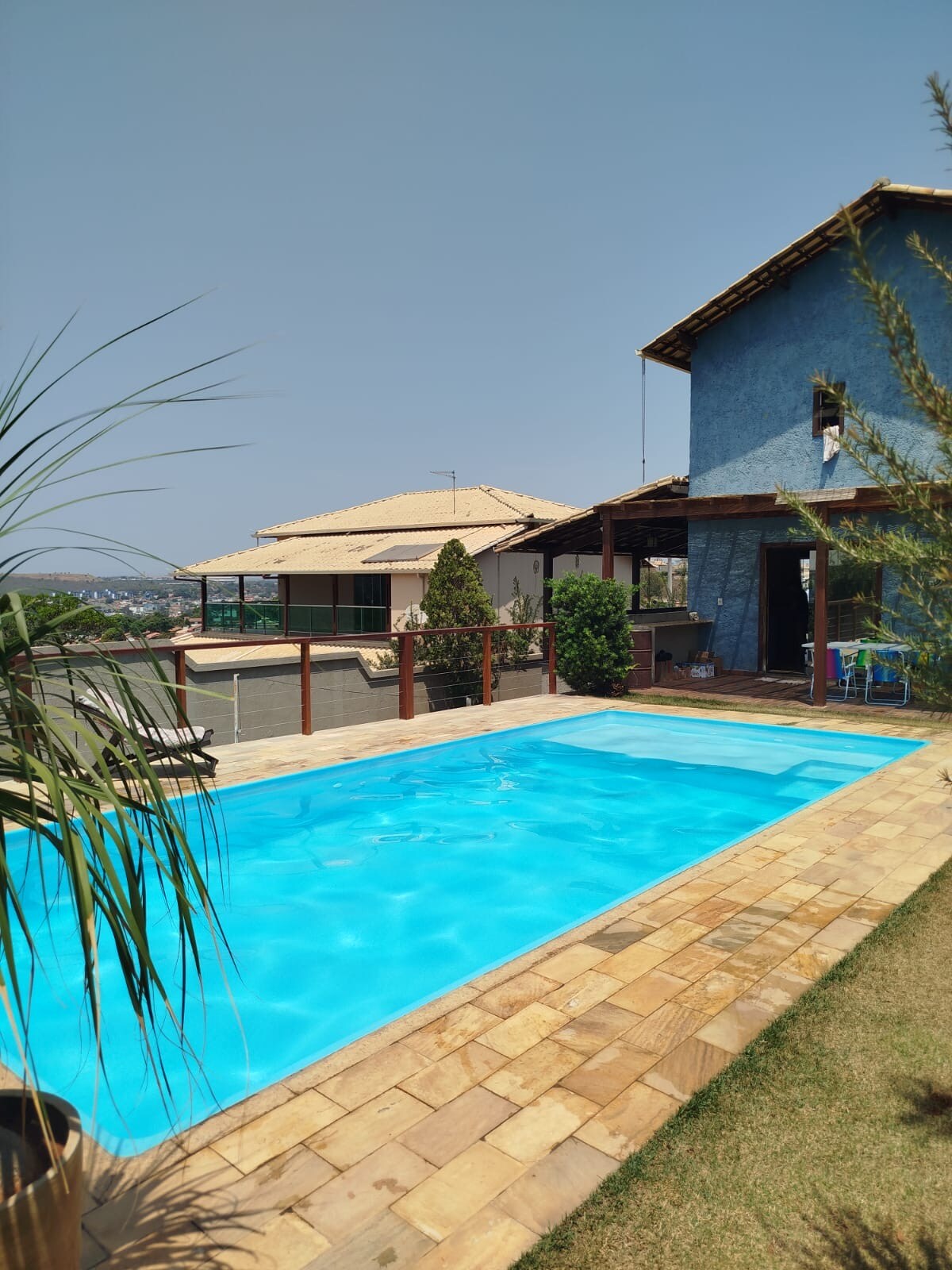 A rectangular pool is surrounded by a wooden deck and lush greenery. The water reflects a clear blue sky, and a two-story blue house is visible in the background. The outdoor area offers a serene space for relaxation.