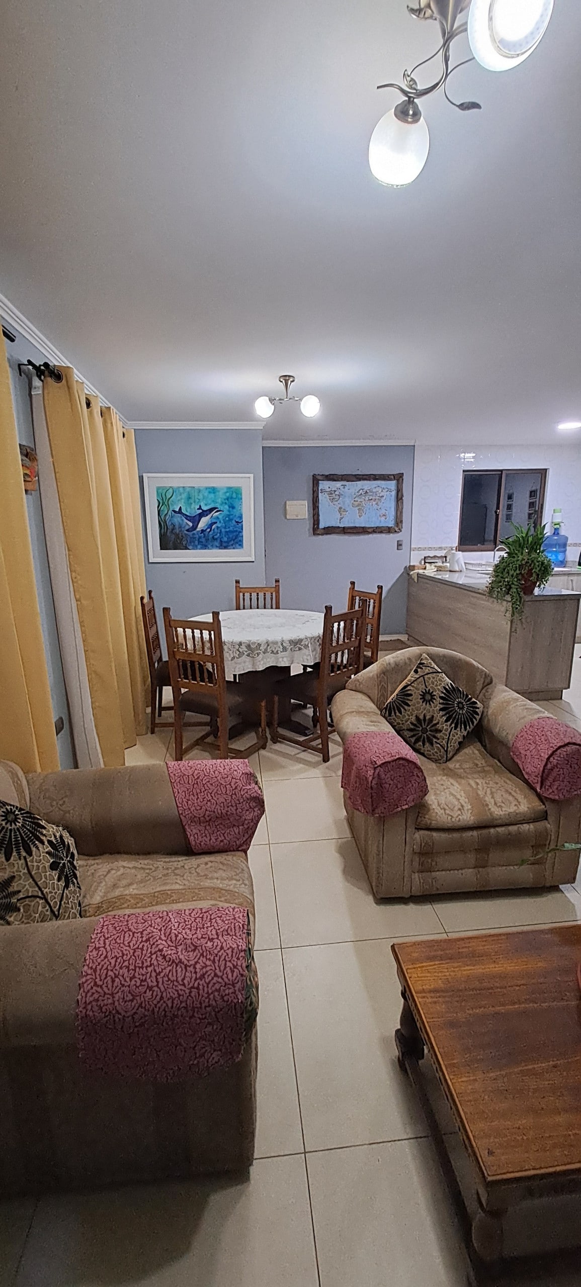 A cozy living area features two armchairs with patterned cushions and a wooden coffee table. A round dining table is surrounded by six wooden chairs, while bright curtains frame the window. The kitchen is partially visible, showcasing modern appliances and a plant in the background.