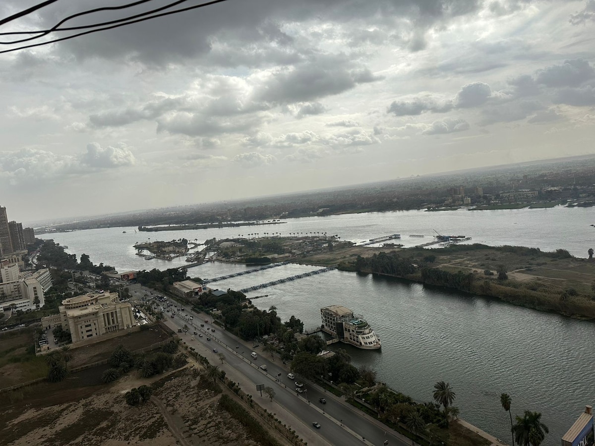 A panoramic view of the Nile River is captured, showcasing a blend of water and greenery along the banks. Streets and vehicles can be seen in the foreground, while distant islands and boats float on the water under an overcast sky.