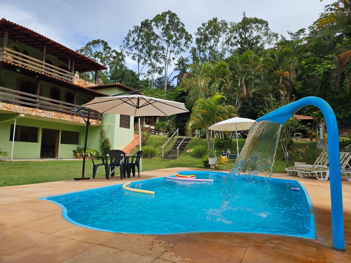 A refreshing pool is situated in a sunny outdoor space, bordered by lush greenery. Several lounge chairs and umbrellas provide shade. A small waterfall feature cascades into the pool, enhancing the overall tranquility of the environment. The house is visible in the background.