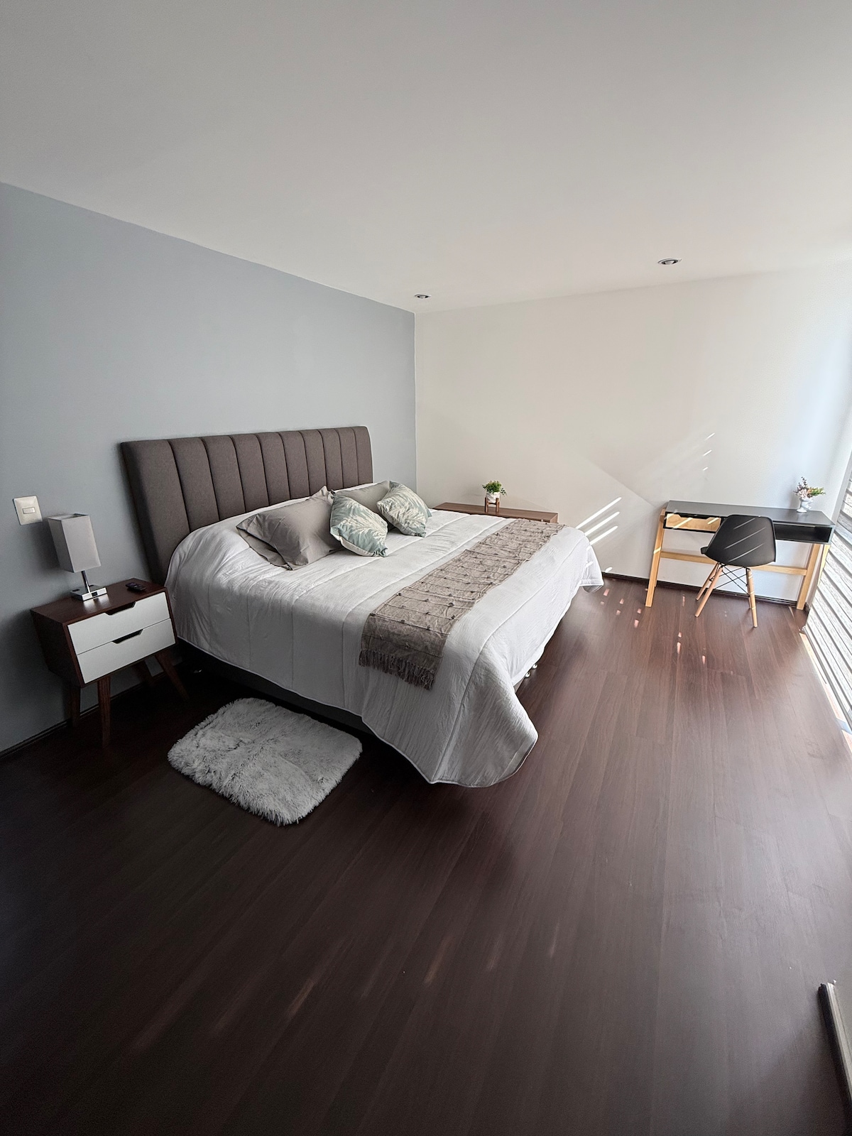 A well-lit bedroom features a spacious bed with a tufted headboard and decorative pillows. A bedside table with a lamp is positioned on each side of the bed. A small desk and chair are arranged near a window, allowing for natural light to fill the space.
