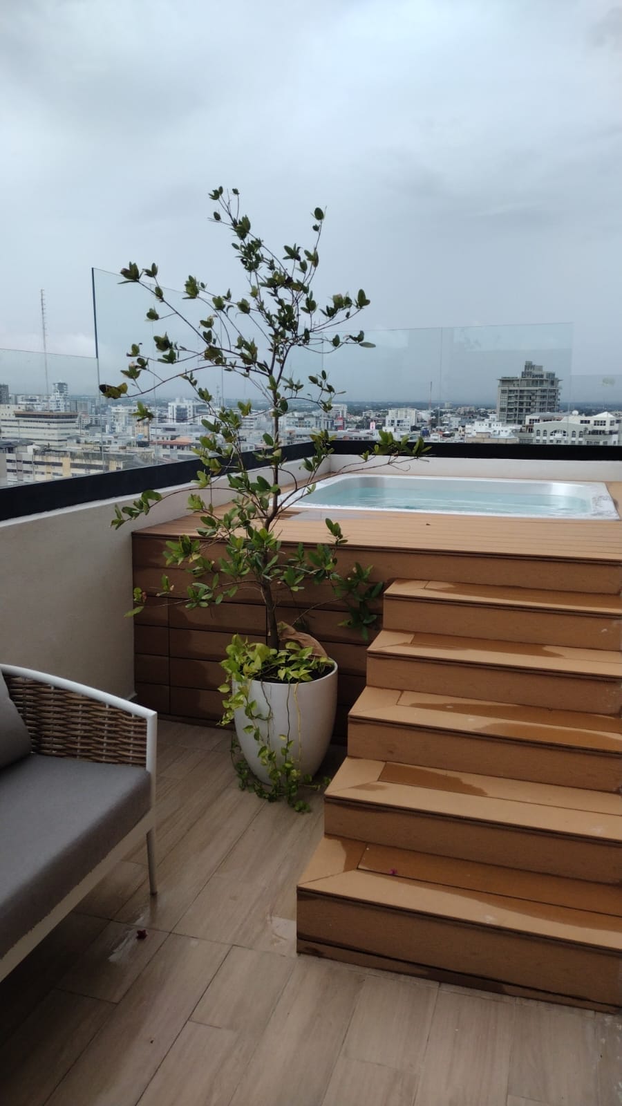An outdoor space is displayed with a small pool surrounded by wooden decking. Steps lead from the pool area to a seating arrangement, featuring a light-colored chair. A tall planter with greenery adds a natural element, enhancing the urban view beyond.