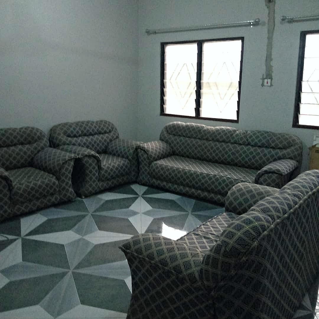 A spacious living area features a patterned sofa set arranged around a geometric tiled floor. Two windows allow natural light to enter, illuminating the room's neutral walls. The furniture is designed for comfort and is ideal for family gatherings.