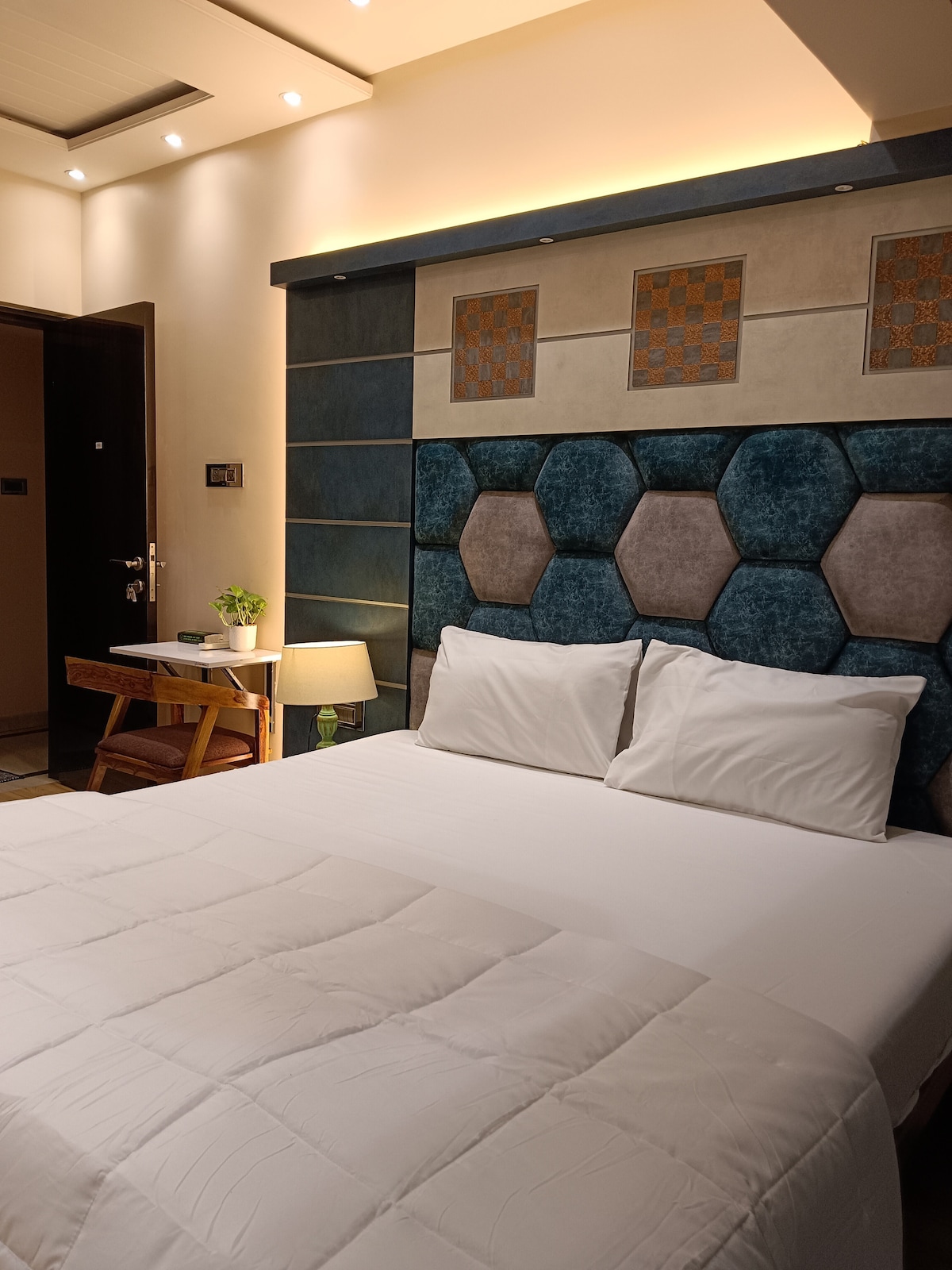 The room features a spacious bed with a plush, hexagon-patterned headboard. A small side table with a lamp is positioned next to the bed. Soft lighting enhances the calming atmosphere, and a potted plant adds a touch of greenery near the entrance.