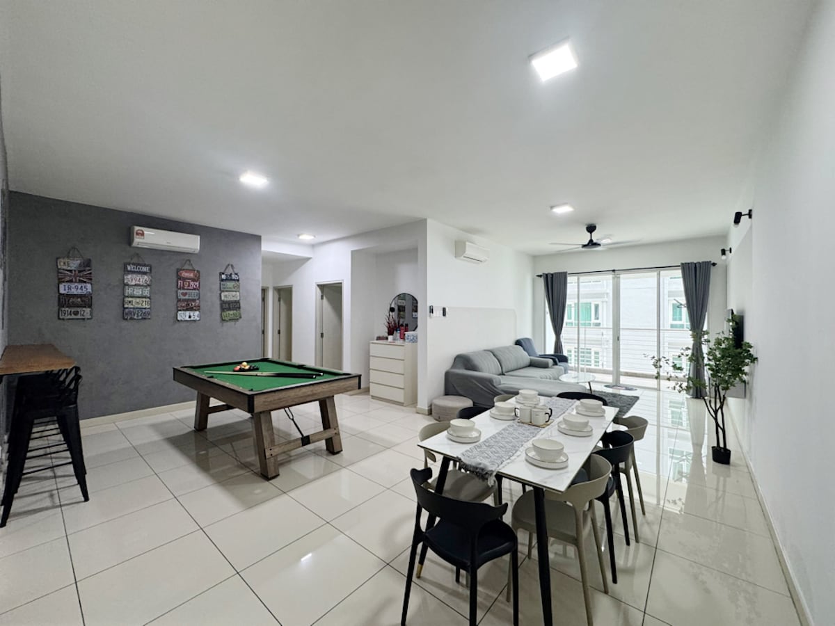 A spacious living area is seen with a large dining table set for meals. A pool table is positioned to one side, and a comfortable seating area with a sofa is visible. Large windows allow natural light to fill the room, enhancing the bright and airy atmosphere.
