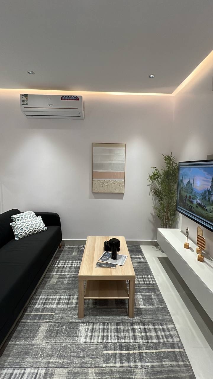 A modern living space features a black sofa accentuated by a patterned pillow. A light wooden coffee table is positioned at the center, holding magazines and decorative items. A large wall-mounted TV displays landscape imagery, while soft ambient lighting enhances the neutral walls.